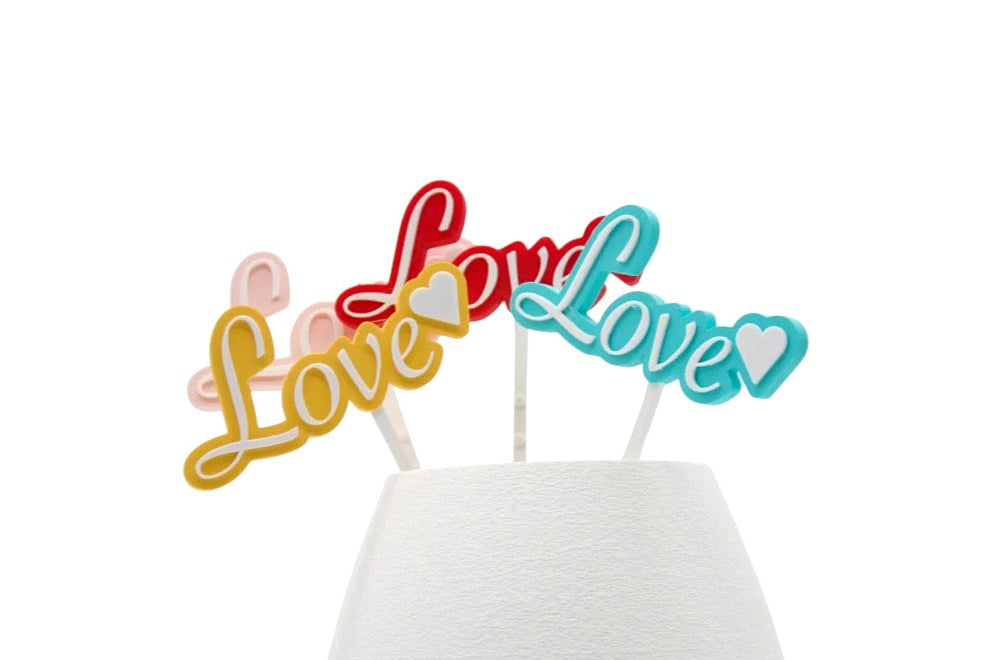 Picks decorativos Love en varios colores colocados sobre base blanca
